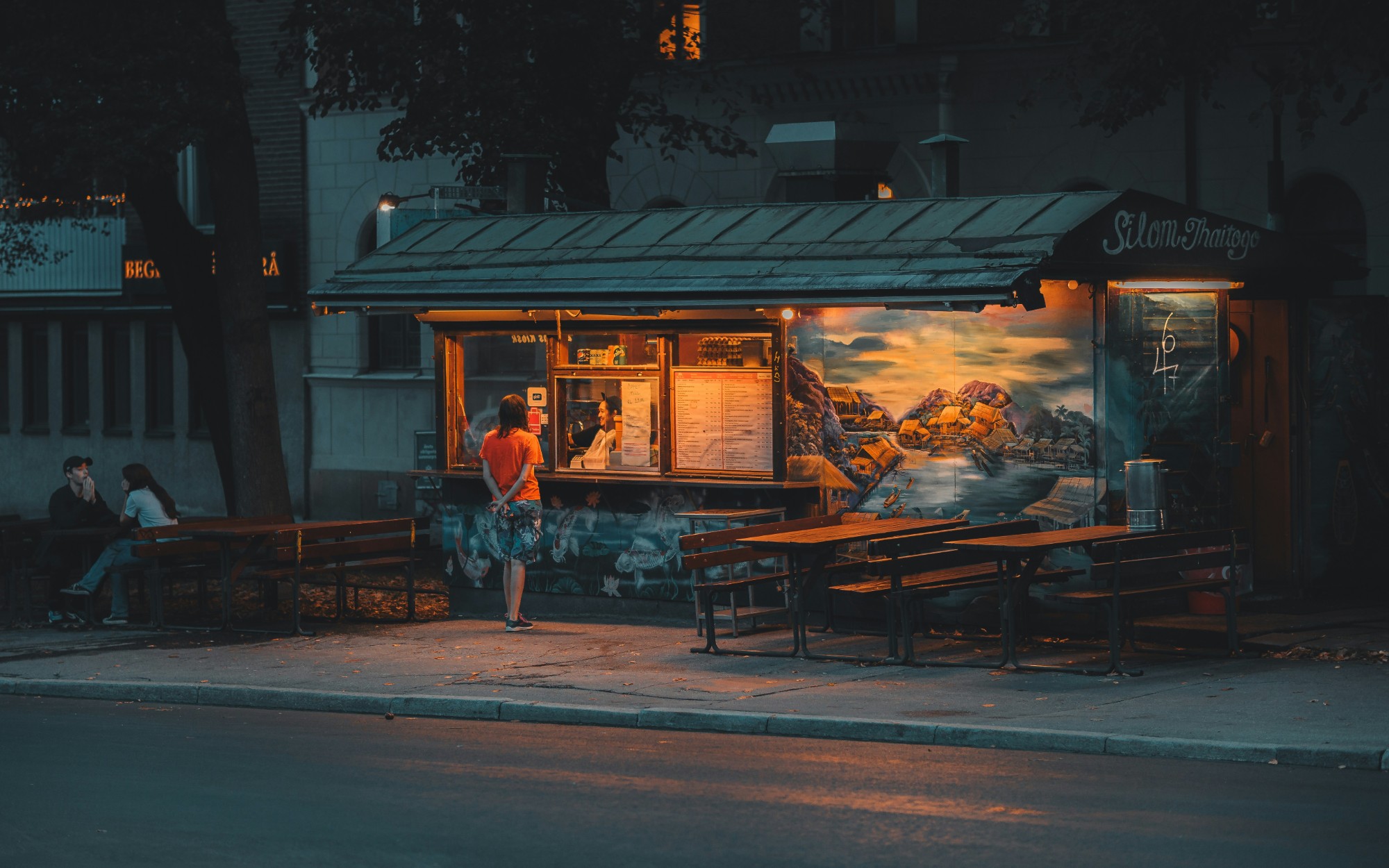 A lit street kiosk at dusk with a painted mural, picnic benches and a person ordering at the window.